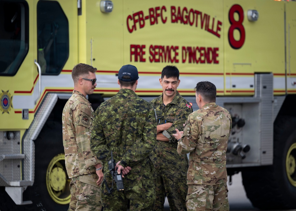 Four military personnel are gathered in conversation in front of a yellow Canadian Forces Base (CFB) Bagotville Fire Service vehicle. The group appears to include both Canadian Armed Forces members and U.S. military personnel in MultiCam/OCP uniforms, suggesting a joint or bilateral military exercise or operation. The setting is an airfield or military base tarmac.