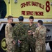 Four military personnel are gathered in conversation in front of a yellow Canadian Forces Base (CFB) Bagotville Fire Service vehicle. The group appears to include both Canadian Armed Forces members and U.S. military personnel in MultiCam/OCP uniforms, suggesting a joint or bilateral military exercise or operation. The setting is an airfield or military base tarmac.