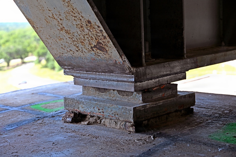 The image shows a close-up view of a steel bridge bearing assembly resting on a concrete pier or abutment cap, photographed from underneath the bridge structure. Significant corrosion and rust are visible on the steel girder and bearing plates, along with deterioration of the concrete base. The background reveals a road and green vegetation, indicating this is an elevated bridge structure.