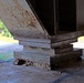 The image shows a close-up view of a steel bridge bearing assembly resting on a concrete pier or abutment cap, photographed from underneath the bridge structure. Significant corrosion and rust are visible on the steel girder and bearing plates, along with deterioration of the concrete base. The background reveals a road and green vegetation, indicating this is an elevated bridge structure.