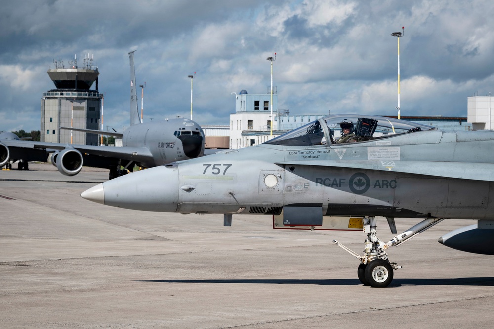 A Royal Canadian Air Force (RCAF) CF-18 Hornet fighter jet numbered '757' is taxiing on an airfield tarmac with a pilot visible in the cockpit. The aircraft bears RCAF markings and the name 'LCol Tremblay-Verrault Seabass' on the fuselage. In the background, a US Air Force large aircraft (likely a KC-135 or similar) and an air traffic control tower are visible.