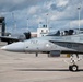 A Royal Canadian Air Force (RCAF) CF-18 Hornet fighter jet numbered '757' is taxiing on an airfield tarmac with a pilot visible in the cockpit. The aircraft bears RCAF markings and the name 'LCol Tremblay-Verrault Seabass' on the fuselage. In the background, a US Air Force large aircraft (likely a KC-135 or similar) and an air traffic control tower are visible.