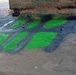 The image shows a close-up of a concrete surface, likely under a bridge or overpass, featuring graffiti spray-painted in bright green and dark grey/black. The graffiti appears to depict a stylized design with curved, leaf-like or flame-like green shapes outlined in dark paint. Additional smaller pink/red text markings are visible in the lower right corner.