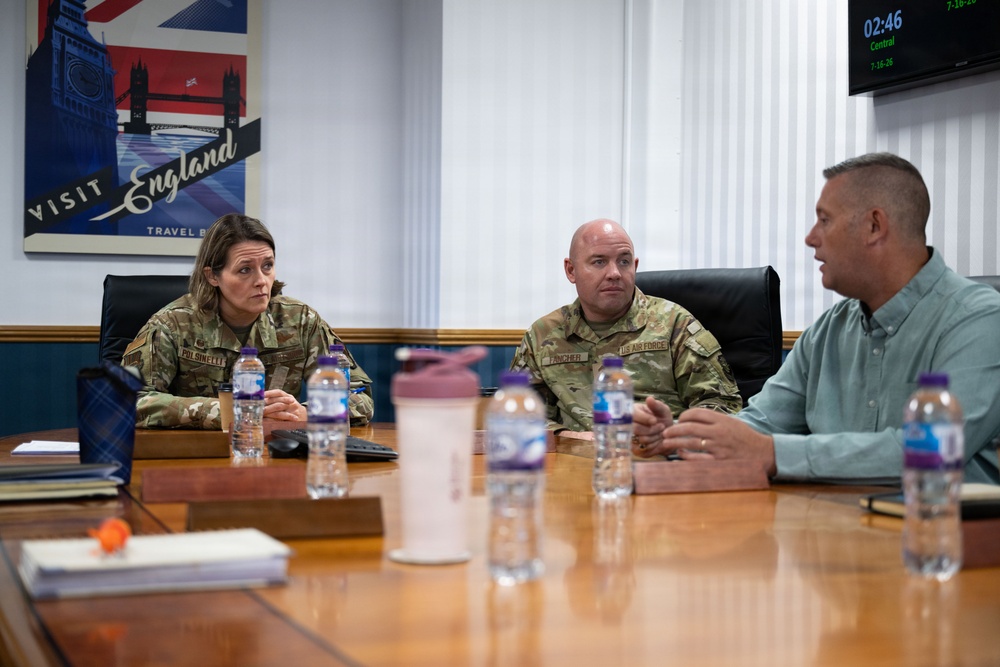 Three individuals are seated at a conference table in what appears to be a military meeting room. Two of the individuals are wearing military camouflage uniforms, while the third is in civilian attire. They appear to be engaged in a formal discussion or briefing.