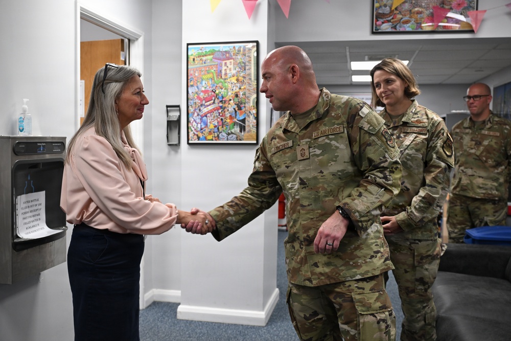 A U.S. Air Force service member is shaking hands with a civilian woman in what appears to be an indoor facility, likely during an official visit or meet-and-greet. Two additional U.S. Air Force personnel in OCP (Operational Camouflage Pattern) uniforms stand in the background. The setting appears to be a community or administrative building decorated with colorful artwork and bunting.