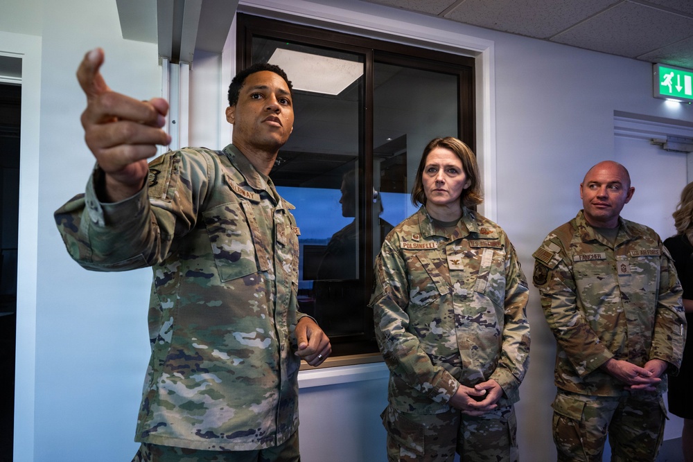 Three U.S. military personnel in OCP (Operational Camouflage Pattern) uniforms are gathered indoors, with one individual gesturing and appearing to brief or explain something to the others. The setting appears to be a military facility or operations building, indicated by the emergency exit sign and interior architecture. Name tapes are partially visible on the uniforms.