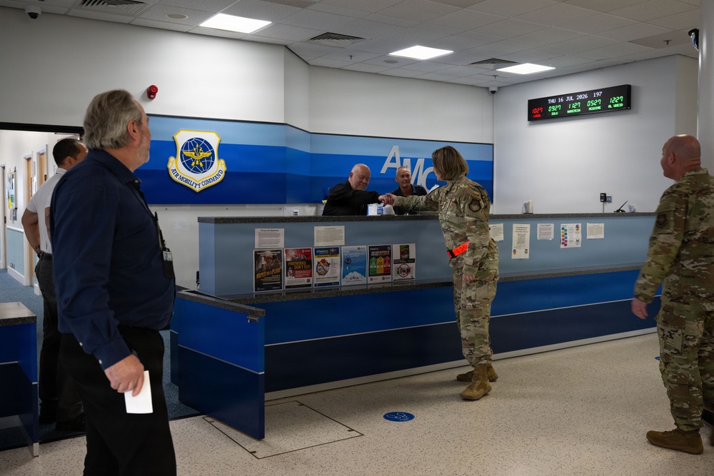 The image shows the interior of an Air Mobility Command (AMC) terminal, likely at a U.S. Air Force base. A female soldier in OCP camouflage uniform is handing documents to staff behind a blue service counter bearing the AMC insignia. Several other individuals, including civilians and at least one other uniformed service member, are present in the facility.