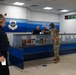 The image shows the interior of an Air Mobility Command (AMC) terminal, likely at a U.S. Air Force base. A female soldier in OCP camouflage uniform is handing documents to staff behind a blue service counter bearing the AMC insignia. Several other individuals, including civilians and at least one other uniformed service member, are present in the facility.