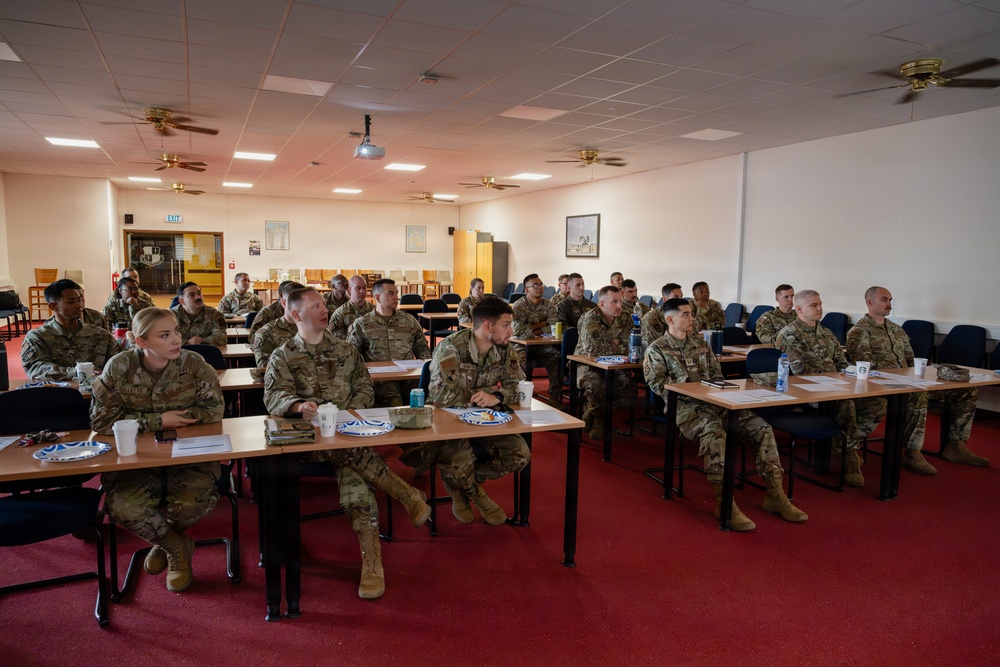A group of approximately 25-30 military personnel in camouflage uniforms (OCP/MultiCam pattern) are seated at desks in a classroom or briefing room setting. They appear to be attending a formal training session or briefing, facing toward a presentation area outside the frame. The room is equipped with a ceiling-mounted projector, ceiling fans, and standard institutional furnishings.