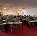 A group of approximately 25-30 military personnel in camouflage uniforms (OCP/MultiCam pattern) are seated at desks in a classroom or briefing room setting. They appear to be attending a formal training session or briefing, facing toward a presentation area outside the frame. The room is equipped with a ceiling-mounted projector, ceiling fans, and standard institutional furnishings.