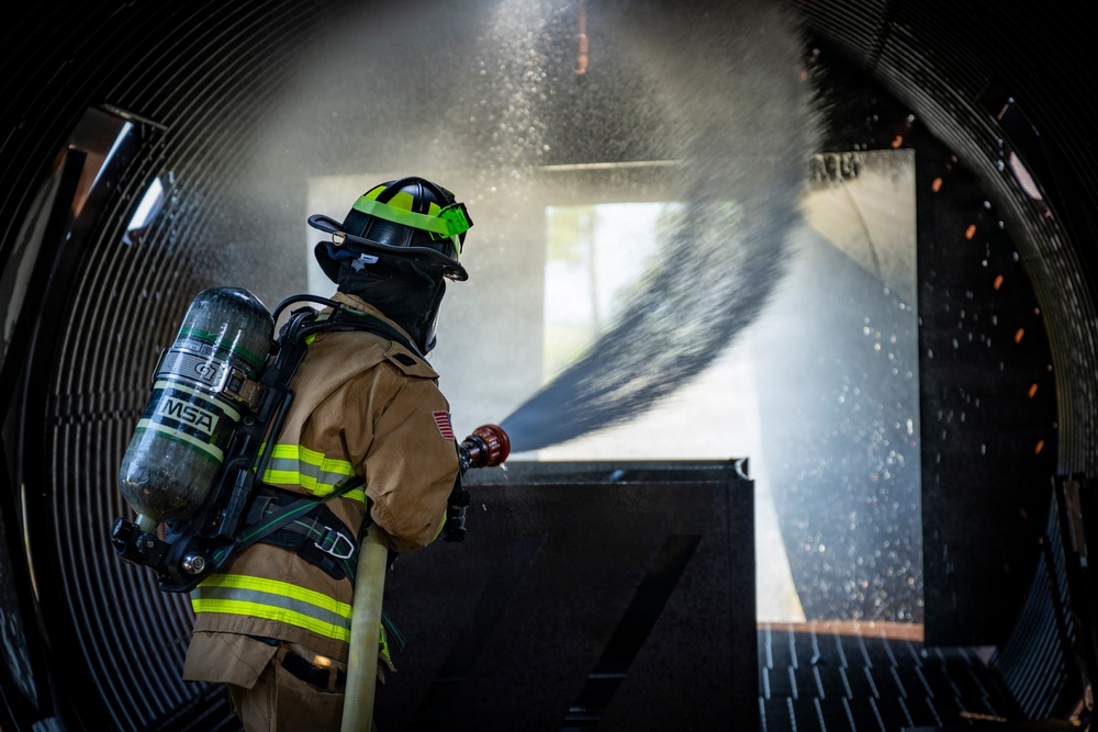 A firefighter in full personal protective equipment is operating a fire hose inside what appears to be a training structure or confined space. The firefighter is directing a water stream forward, with visible water spray and mist filling the environment. The image captures a likely live-fire training or suppression exercise scenario.