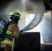 A firefighter in full personal protective equipment is operating a fire hose inside what appears to be a training structure or confined space. The firefighter is directing a water stream forward, with visible water spray and mist filling the environment. The image captures a likely live-fire training or suppression exercise scenario.
