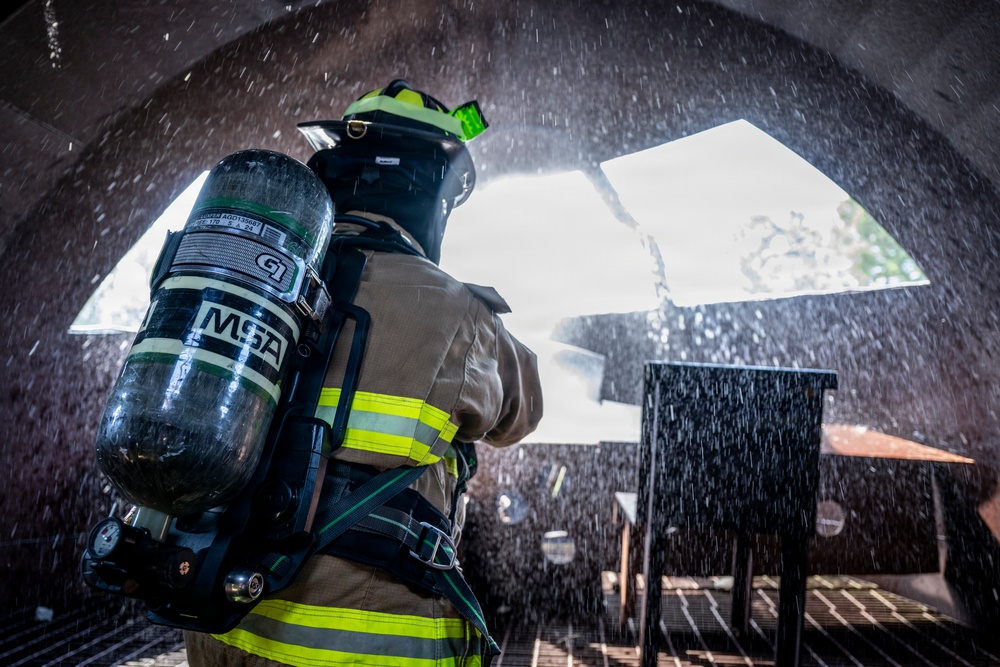 A firefighter in full turnout gear is seen from behind, carrying an MSA self-contained breathing apparatus (SCBA) tank, standing beneath an arched opening with water spray visible throughout the scene. The setting appears to be a training exercise or active firefighting operation, with water droplets captured mid-air suggesting a hose or sprinkler system in use. The low-angle perspective emphasizes the SCBA equipment and the firefighter's protective gear.