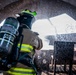 A firefighter in full turnout gear is seen from behind, carrying an MSA self-contained breathing apparatus (SCBA) tank, standing beneath an arched opening with water spray visible throughout the scene. The setting appears to be a training exercise or active firefighting operation, with water droplets captured mid-air suggesting a hose or sprinkler system in use. The low-angle perspective emphasizes the SCBA equipment and the firefighter's protective gear.