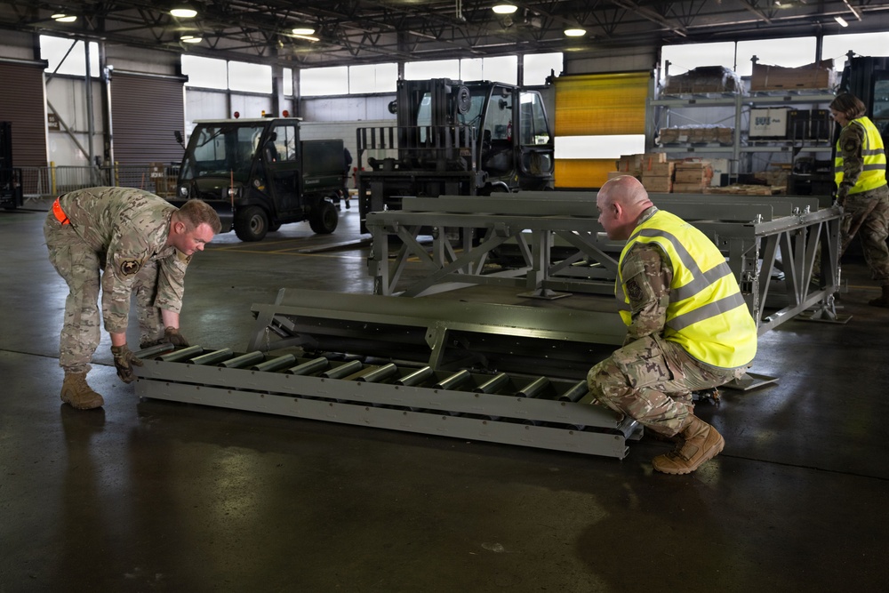 Military personnel in MultiCam/MTP camouflage uniforms are working inside a large warehouse or hangar, handling what appears to be aircraft cargo loading equipment with roller conveyors. Two soldiers in the foreground are positioning or inspecting a roller conveyor pallet system, while a third person works in the background. The facility contains forklifts, utility vehicles, and stacked wooden crates, indicating an active logistics or air cargo operation.