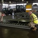 Military personnel in MultiCam/MTP camouflage uniforms are working inside a large warehouse or hangar, handling what appears to be aircraft cargo loading equipment with roller conveyors. Two soldiers in the foreground are positioning or inspecting a roller conveyor pallet system, while a third person works in the background. The facility contains forklifts, utility vehicles, and stacked wooden crates, indicating an active logistics or air cargo operation.