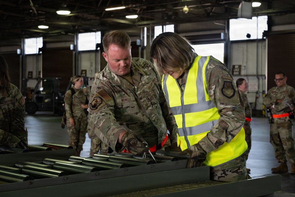 Two U.S. Air Force personnel in OCP (Operational Camouflage Pattern) uniforms are working together at a table with cylindrical metal components in what appears to be a large hangar or warehouse facility. The individual on the right is wearing a high-visibility safety vest over their uniform, suggesting a supervisory or safety role. Several other uniformed personnel are visible in the background observing or participating in the activity.