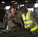 Two U.S. Air Force personnel in OCP (Operational Camouflage Pattern) uniforms are working together at a table with cylindrical metal components in what appears to be a large hangar or warehouse facility. The individual on the right is wearing a high-visibility safety vest over their uniform, suggesting a supervisory or safety role. Several other uniformed personnel are visible in the background observing or participating in the activity.