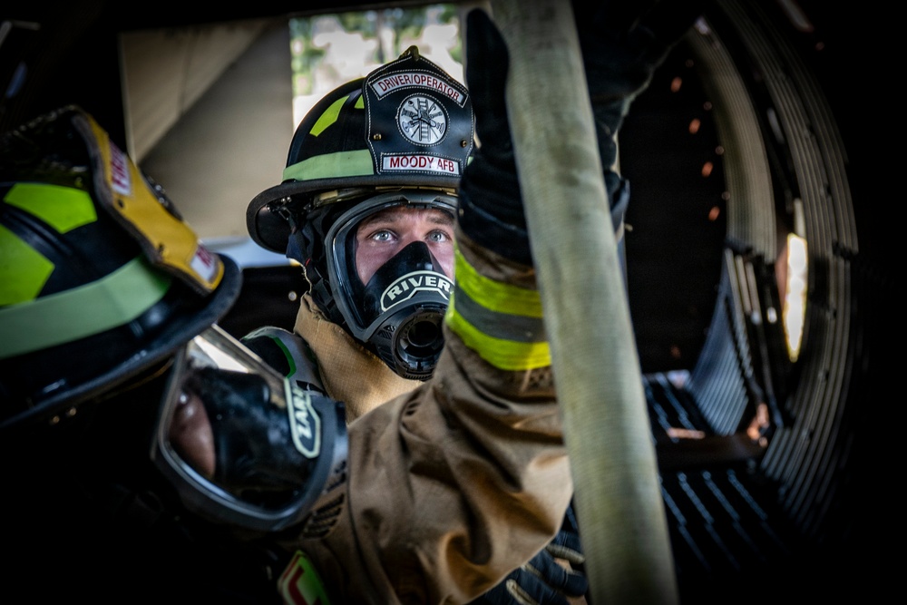 A firefighter in full turnout gear is photographed in close-up, wearing a black helmet labeled 'MOODY AFB' and 'DRIVER/OPERATOR' with a breathing mask marked 'RIVER'. The subject appears to be positioned near or on a fire apparatus, with a large hose reel visible in the background. The image is a professional, high-contrast photograph likely taken at Moody Air Force Base.
