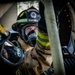 A firefighter in full turnout gear is photographed in close-up, wearing a black helmet labeled 'MOODY AFB' and 'DRIVER/OPERATOR' with a breathing mask marked 'RIVER'. The subject appears to be positioned near or on a fire apparatus, with a large hose reel visible in the background. The image is a professional, high-contrast photograph likely taken at Moody Air Force Base.