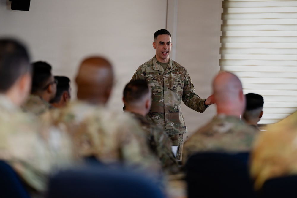 A military service member in OCP (Operational Camouflage Pattern) uniform is addressing a group of fellow service members in what appears to be a briefing or training session. The speaker is standing at the front of the room while the audience is seated, viewed from behind in soft focus. The setting appears to be an indoor meeting or conference room on a military installation.