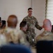 A military service member in OCP (Operational Camouflage Pattern) uniform is addressing a group of fellow service members in what appears to be a briefing or training session. The speaker is standing at the front of the room while the audience is seated, viewed from behind in soft focus. The setting appears to be an indoor meeting or conference room on a military installation.