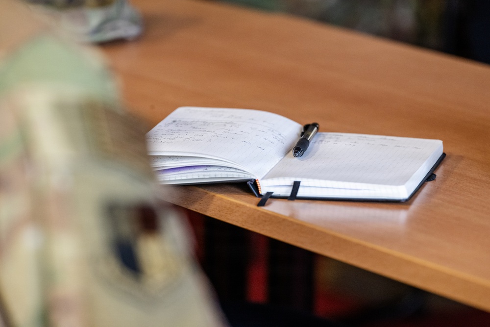 An open notebook with handwritten notes lies on a wooden desk, with a black pen resting across its pages. The notebook appears well-used, with many filled pages visible on the left side and a cleaner right page. A bookmark ribbon is visible along the notebook's edge.