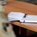 An open notebook with handwritten notes lies on a wooden desk, with a black pen resting across its pages. The notebook appears well-used, with many filled pages visible on the left side and a cleaner right page. A bookmark ribbon is visible along the notebook's edge.