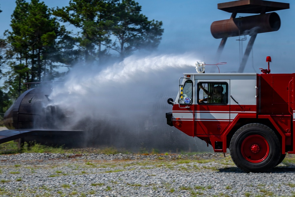 An airport rescue and firefighting (ARFF) vehicle, painted red and white, is actively discharging a powerful water stream from its roof-mounted turret nozzle toward what appears to be a mock aircraft fuselage used for training exercises. The scene takes place on a gravel surface with trees visible in the background, suggesting a dedicated fire training area. A operator is visible inside the cab controlling the vehicle during what appears to be a live fire suppression drill.
