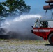 An airport rescue and firefighting (ARFF) vehicle, painted red and white, is actively discharging a powerful water stream from its roof-mounted turret nozzle toward what appears to be a mock aircraft fuselage used for training exercises. The scene takes place on a gravel surface with trees visible in the background, suggesting a dedicated fire training area. A operator is visible inside the cab controlling the vehicle during what appears to be a live fire suppression drill.