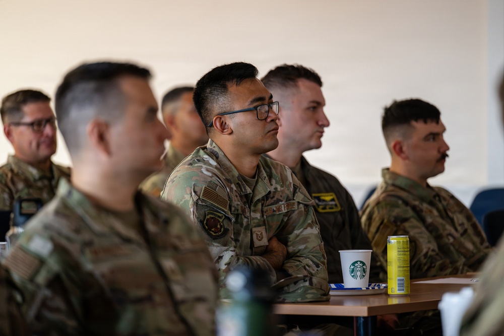 A group of U.S. Air Force personnel in OCP (Operational Camouflage Pattern) uniforms are seated and attentively listening to a briefing or presentation. The central figure, wearing glasses, has his arms crossed and appears focused on something outside the frame. The setting appears to be an indoor briefing room or classroom environment.