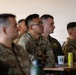 A group of U.S. Air Force personnel in OCP (Operational Camouflage Pattern) uniforms are seated and attentively listening to a briefing or presentation. The central figure, wearing glasses, has his arms crossed and appears focused on something outside the frame. The setting appears to be an indoor briefing room or classroom environment.