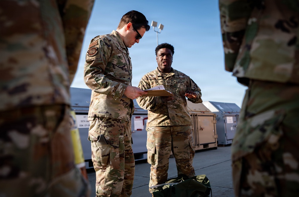 Two military personnel in OCP (Operational Camouflage Pattern) uniforms are engaged in a discussion outdoors, with one reviewing a document or card while the other gestures and speaks. They appear to be on a military installation or flight line, surrounded by equipment containers and storage units. A duffel bag is visible on the ground between them.