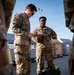Two military personnel in OCP (Operational Camouflage Pattern) uniforms are engaged in a discussion outdoors, with one reviewing a document or card while the other gestures and speaks. They appear to be on a military installation or flight line, surrounded by equipment containers and storage units. A duffel bag is visible on the ground between them.