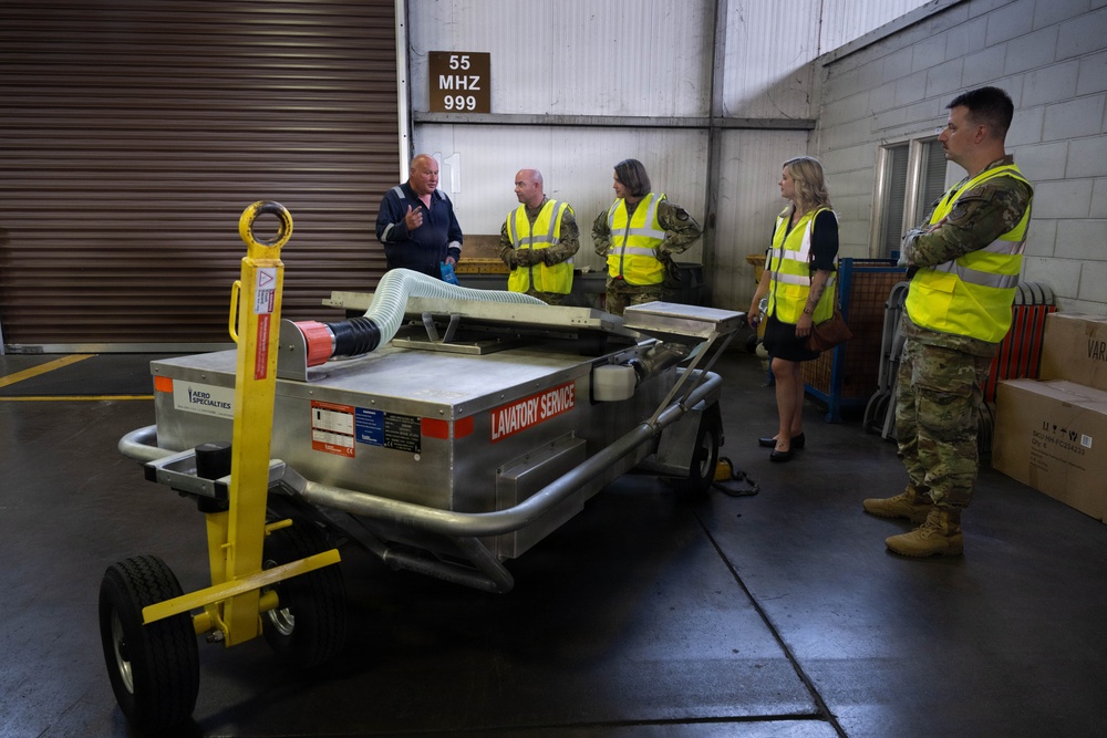 A group of individuals in a warehouse or hangar facility are gathered around an aircraft lavatory service cart manufactured by Aero Specialties. The group includes two military personnel in camouflage uniforms, two civilians wearing high-visibility yellow safety vests, and a presenter in a blue uniform who appears to be conducting a briefing or demonstration. A sign on the wall reads '55 MHZ 999', suggesting a military or aviation facility.