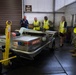 A group of individuals in a warehouse or hangar facility are gathered around an aircraft lavatory service cart manufactured by Aero Specialties. The group includes two military personnel in camouflage uniforms, two civilians wearing high-visibility yellow safety vests, and a presenter in a blue uniform who appears to be conducting a briefing or demonstration. A sign on the wall reads '55 MHZ 999', suggesting a military or aviation facility.