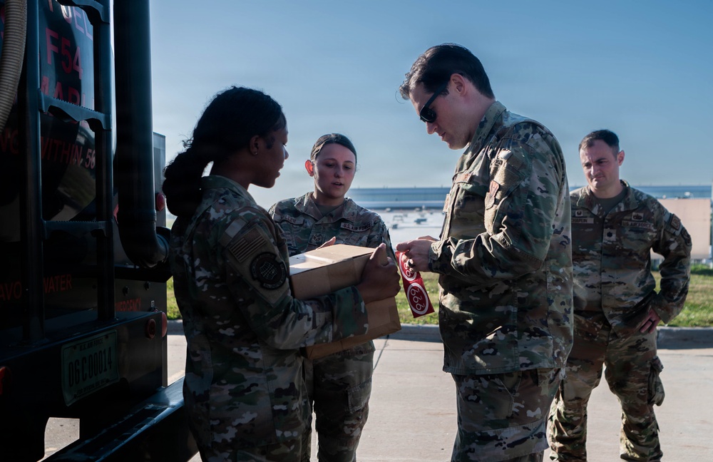 Four military personnel in OCP (Operational Camouflage Pattern) uniforms are gathered near a military vehicle outdoors. A senior officer appears to be examining or signing a red tag/label attached to a cardboard box being held by a female service member. Two additional personnel observe the interaction in the background.