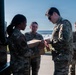 Four military personnel in OCP (Operational Camouflage Pattern) uniforms are gathered near a military vehicle outdoors. A senior officer appears to be examining or signing a red tag/label attached to a cardboard box being held by a female service member. Two additional personnel observe the interaction in the background.