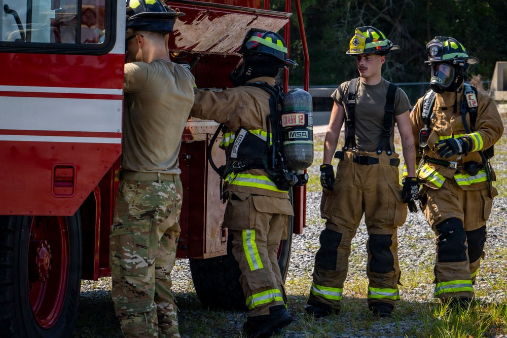 The image shows a group of military firefighters and personnel gathered around a red fire apparatus vehicle, likely during a training exercise. Two individuals are wearing full firefighting turnout gear with SCBA (Self-Contained Breathing Apparatus) units, while others are in military camouflage uniforms. The setting appears to be an outdoor training area with gravel ground.