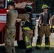 The image shows a group of military firefighters and personnel gathered around a red fire apparatus vehicle, likely during a training exercise. Two individuals are wearing full firefighting turnout gear with SCBA (Self-Contained Breathing Apparatus) units, while others are in military camouflage uniforms. The setting appears to be an outdoor training area with gravel ground.