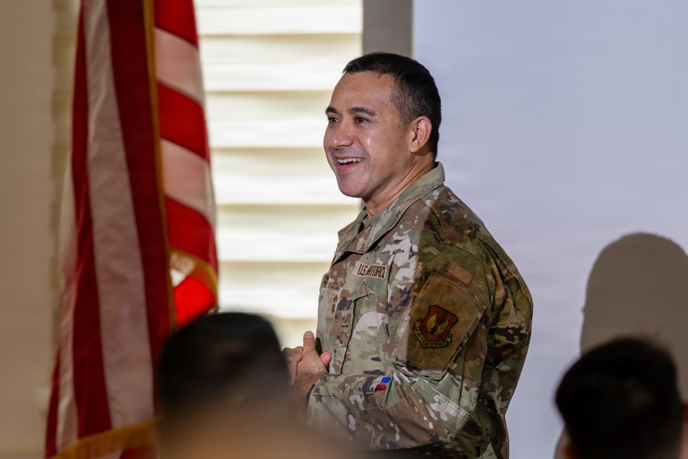 A U.S. military service member in OCP (Operational Camouflage Pattern) uniform is smiling and appears to be speaking or presenting to an audience. An American flag on a ceremonial pole is visible to his left. The setting appears to be an indoor military ceremony or briefing.