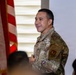 A U.S. military service member in OCP (Operational Camouflage Pattern) uniform is smiling and appears to be speaking or presenting to an audience. An American flag on a ceremonial pole is visible to his left. The setting appears to be an indoor military ceremony or briefing.