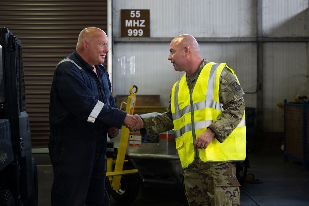Two men are shaking hands inside what appears to be a military or industrial facility. One man is wearing dark blue coveralls while the other is dressed in military camouflage uniform with a high-visibility yellow safety vest. The setting suggests a formal or semi-formal greeting in a maintenance or logistics environment.