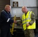 Two men are shaking hands inside what appears to be a military or industrial facility. One man is wearing dark blue coveralls while the other is dressed in military camouflage uniform with a high-visibility yellow safety vest. The setting suggests a formal or semi-formal greeting in a maintenance or logistics environment.