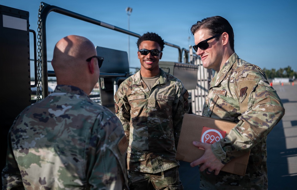 Three U.S. Air Force personnel in OCP (Operational Camouflage Pattern) uniforms are engaged in conversation outdoors on what appears to be a flight line or military installation. One airman is holding a cardboard box with a red circular logo, while the others face him in discussion. The setting appears to be a sunny day with military vehicles visible in the background.