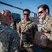 Three U.S. Air Force personnel in OCP (Operational Camouflage Pattern) uniforms are engaged in conversation outdoors on what appears to be a flight line or military installation. One airman is holding a cardboard box with a red circular logo, while the others face him in discussion. The setting appears to be a sunny day with military vehicles visible in the background.