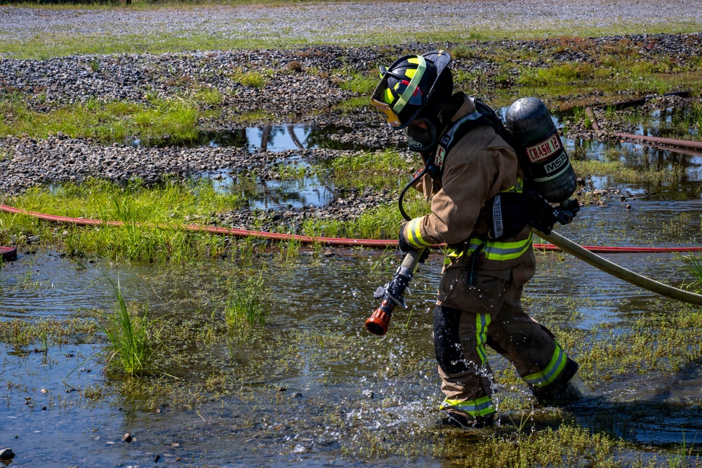 A firefighter in full personal protective equipment is wading through shallow water while dragging fire hoses across a wet, marshy terrain. The firefighter is wearing a self-contained breathing apparatus (SCBA) and tan turnout gear with high-visibility yellow reflective striping. This appears to be a training exercise or active emergency response in an outdoor, waterlogged environment.