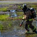 A firefighter in full personal protective equipment is wading through shallow water while dragging fire hoses across a wet, marshy terrain. The firefighter is wearing a self-contained breathing apparatus (SCBA) and tan turnout gear with high-visibility yellow reflective striping. This appears to be a training exercise or active emergency response in an outdoor, waterlogged environment.