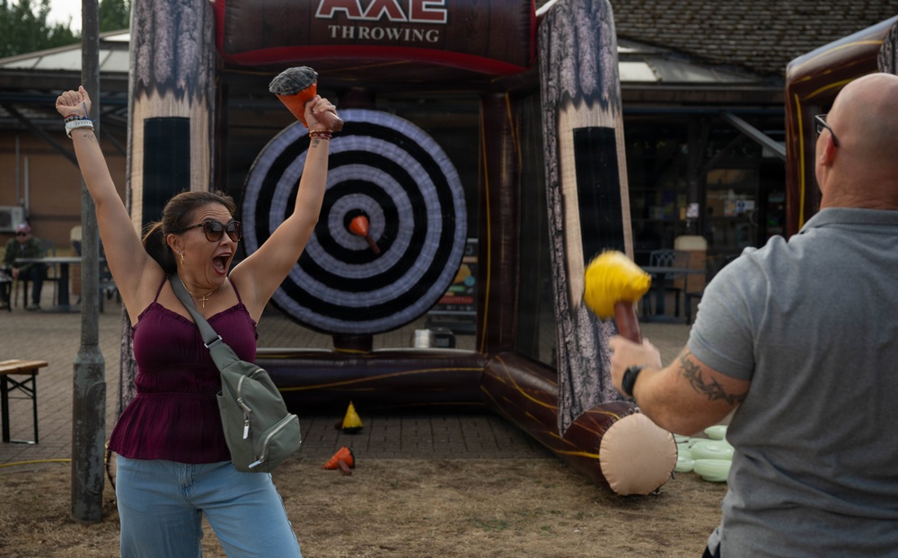 Two people are participating in an inflatable axe throwing game at what appears to be an outdoor fair or event. A woman in a maroon top is celebrating enthusiastically with her arms raised, holding a foam/rubber axe, while a bald man with tattoos faces the target. An orange foam axe is visibly stuck near the bullseye of the inflatable target.