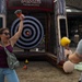 Two people are participating in an inflatable axe throwing game at what appears to be an outdoor fair or event. A woman in a maroon top is celebrating enthusiastically with her arms raised, holding a foam/rubber axe, while a bald man with tattoos faces the target. An orange foam axe is visibly stuck near the bullseye of the inflatable target.
